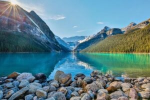 Tranquil morning view of Lake Louise in the Canadian Rockies with sun rays peeking over the mountains and reflecting in turquoise waters.