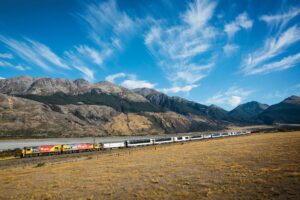KiwiRail’s TranzAlpine train journeying across golden plains beneath dramatic mountain peaks in New Zealand.