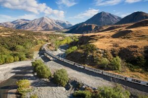 The TranzAlpine train crossing the Cass River and Mt White Bridge surrounded by South Island’s mountainous terrain.