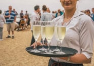 Smiling server holding a tray of chilled sparkling wine glasses with beachside guests in the background.